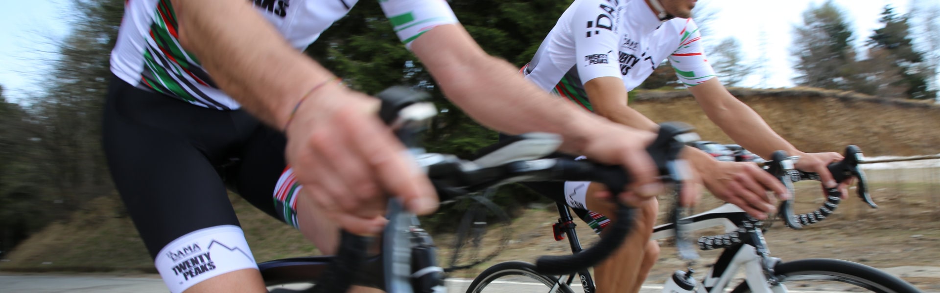 Two road cyclists in Twenty Peaks kit riding side by side along a tree-lined road